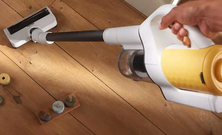 Un aspirateur blanc et jaune nettoie un plancher de bois. Des jouets en bois colorés sont visibles sur le plancher.