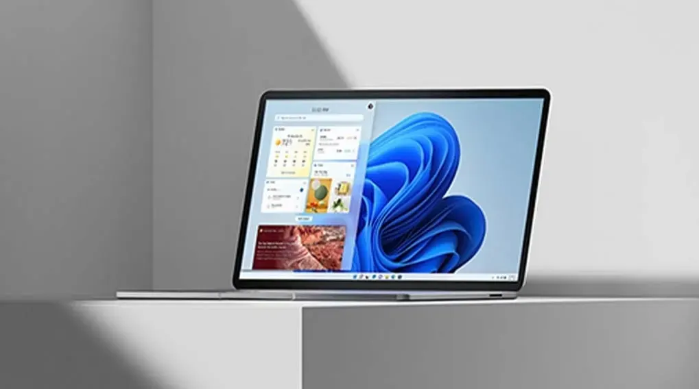 Zilverkleurige laptop op witte ondergrond. Het scherm toont een blauw abstract patroon en een widget met weerinformatie.
