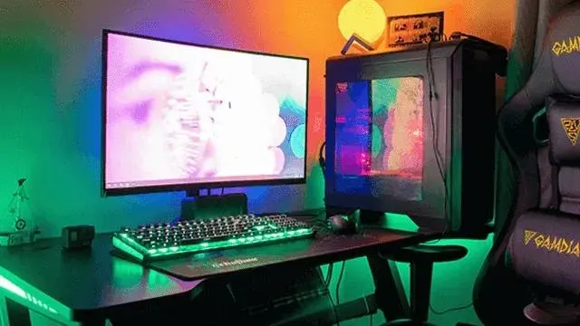Bureau avec écran, clavier éclairé, unité centrale, casque et chaise gamer noire. Éclairage ambiant coloré.