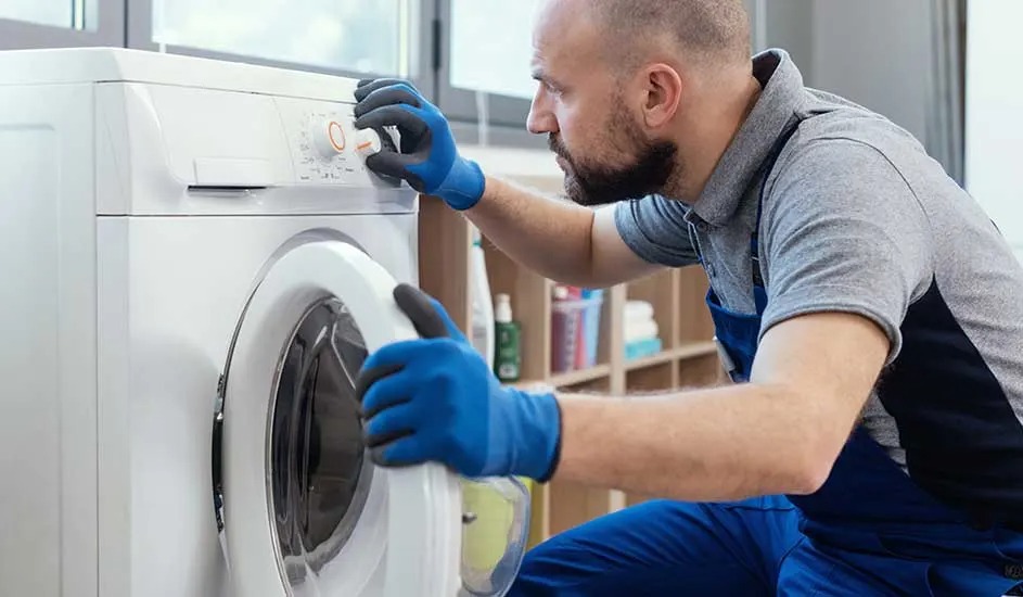 Een man met blauwe handschoenen bedient het paneel van een witte wasmachine en houdt de deur open, merk is niet zichtbaar.