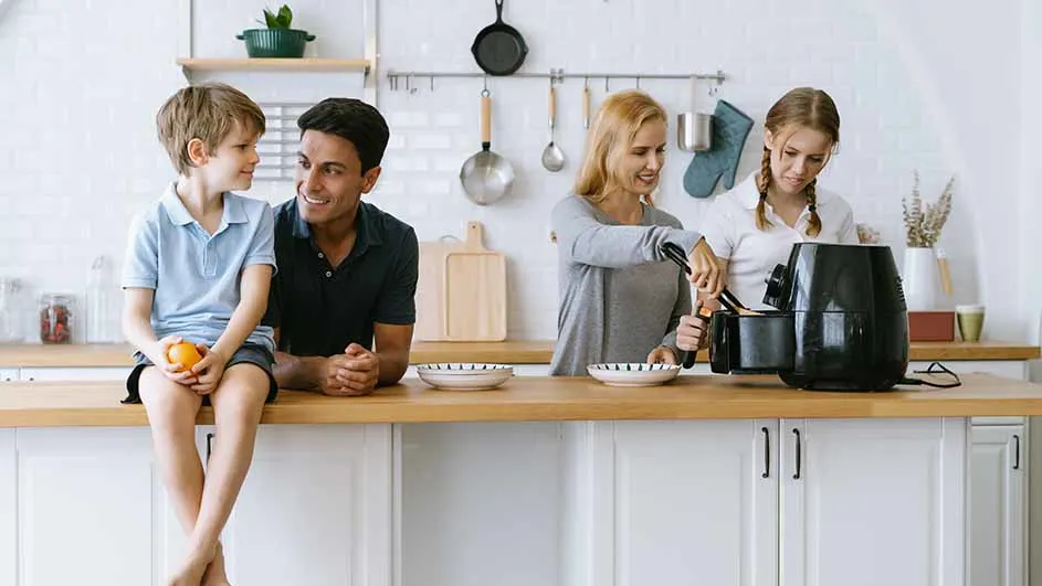 In een keuken staat een zwarte airfryer op een houten aanrecht. Borden staan klaar. Keukengerei hangt aan de muur.
