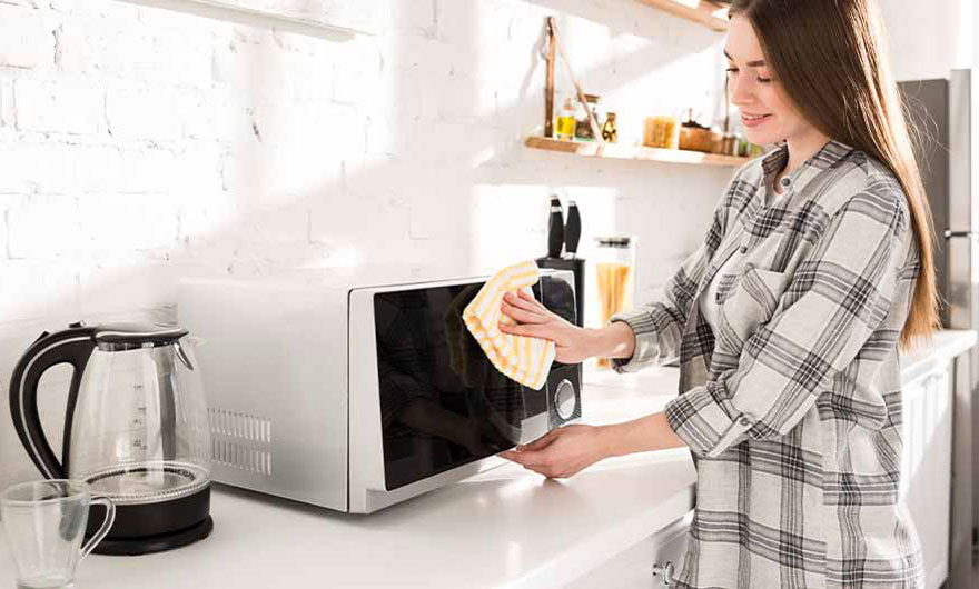 Une cuisine lumineuse avec un micro-ondes blanc nettoyé avec un chiffon jaune, près d'une bouilloire en verre.