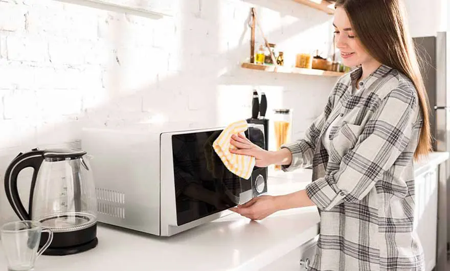 Une cuisine lumineuse avec un micro-ondes blanc nettoyé avec un chiffon jaune, près d'une bouilloire en verre.
