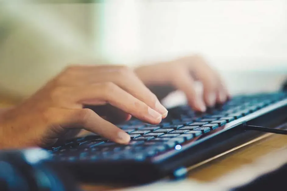 Mains tapant sur un clavier noir sur un bureau en bois clair. L'arrière-plan est clair.