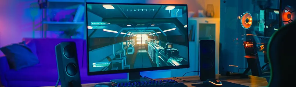 A gaming setup with a monitor showing a first-person shooter game, keyboard, mouse, speakers, and a lit-up computer tower.