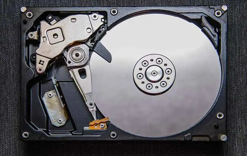 Een geopende harde schijf met zilveren schijf en metalen leesarm, tegen een donkergrijze achtergrond.