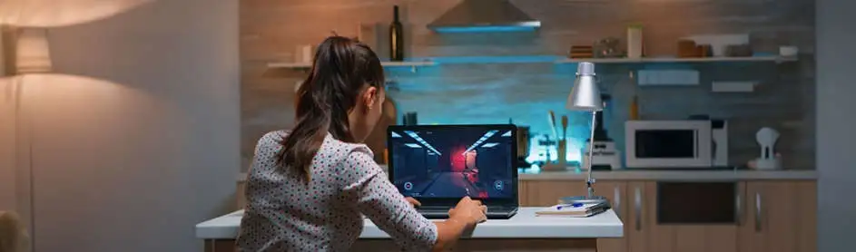 A laptop displays a video game on a white table with a silver lamp. Kitchen cabinets and a microwave are in the background.