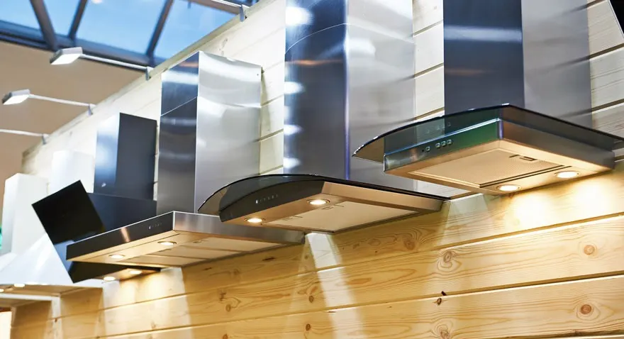 Rangée de hottes de cuisine en acier inoxydable et noires montées sur un mur en bois clair. Luminaires visibles au plafond.