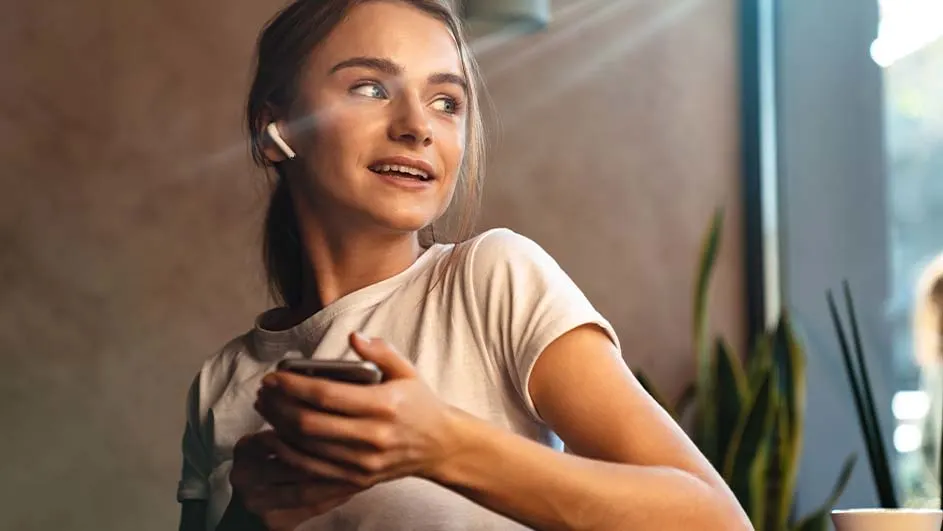 Une personne avec un téléphone et des écouteurs. À l'arrière-plan, on voit des plantes et une fenêtre.