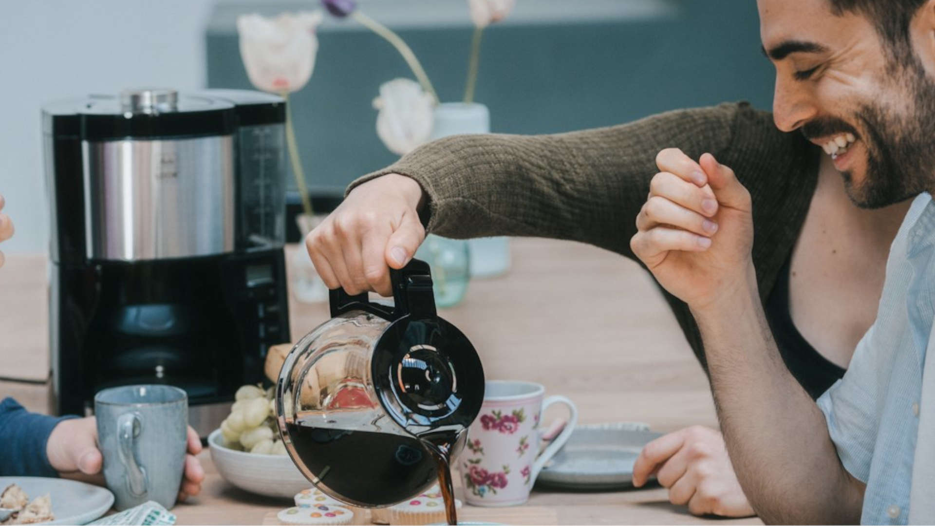 A coffee maker is on the table next to a jug from which coffee is being poured into a cup.