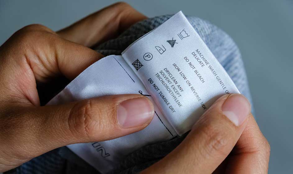 Handen houden een wit kledinglabel vast bij een grijze stof, met wasinstructies, kleine symbolen en tekst duidelijk in beeld.