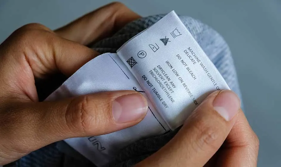 Handen houden een wit kledinglabel vast bij een grijze stof, met wasinstructies, kleine symbolen en tekst duidelijk in beeld.