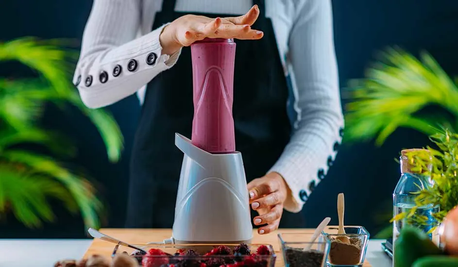 Préparation d'un smoothie : mixer blanc et récipient rose sur table en bois, avec fruits rouges, herbes et bouteille en arrière-plan.