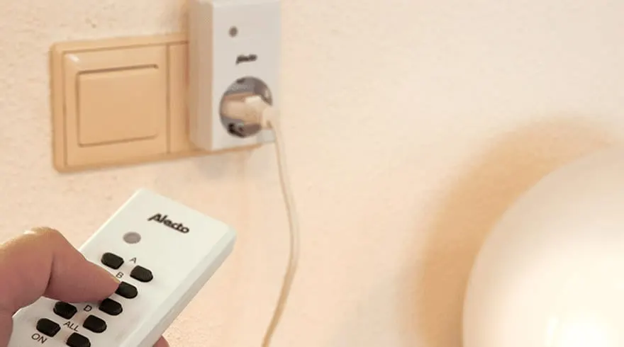 Une télécommande blanche Alecto est pointée vers une prise murale blanche. Un interrupteur beige est visible.