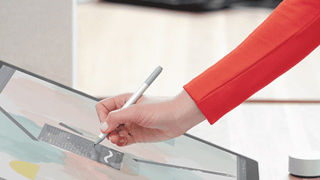 Une main en veste rouge utilise un stylet sur une tablette grise, un écran affiche une règle, sur un fond blanc.