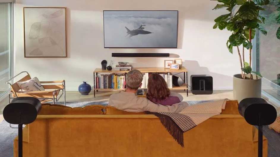 Woonkamer met mosterdkleurige bank, tv met vliegtuigbeeld, houten stoel, plant en boekenkast tegen een witte muur.