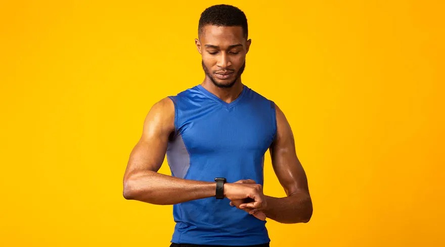 Een man met een blauw hemd kijkt naar een zwarte smartwatch tegen een gele achtergrond.