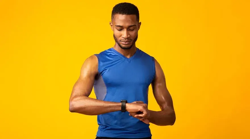 Een man met een blauw hemd kijkt naar een zwarte smartwatch tegen een gele achtergrond.