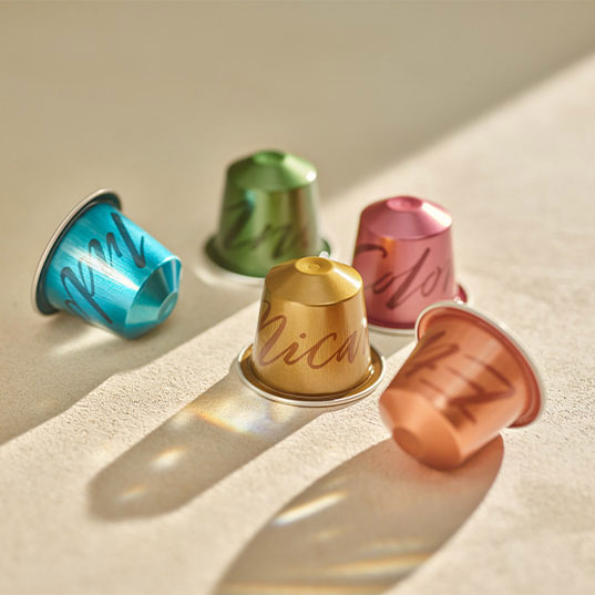 Vijf kleurrijke koffiecapsules (blauw, groen, goud, roze en koper) liggen op een licht oppervlak met schaduwen.