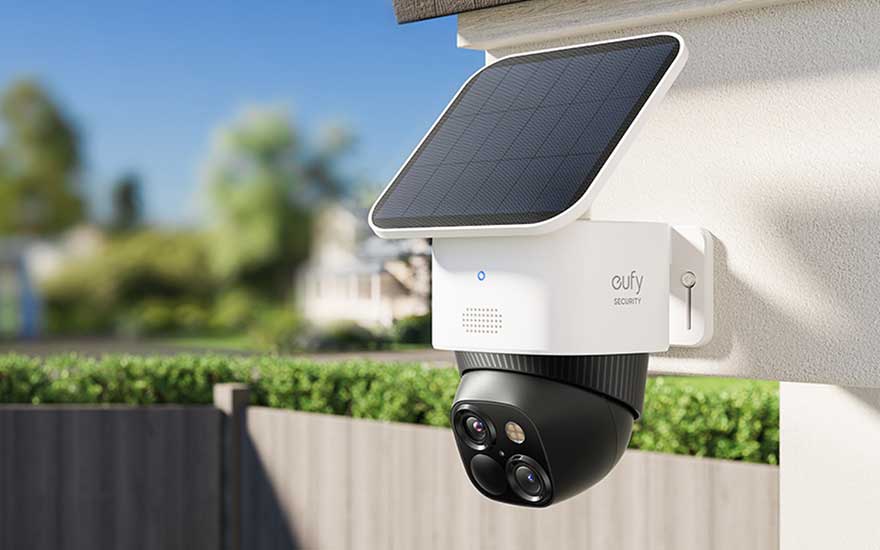 Caméra de sécurité blanche et noire avec panneau solaire sur un mur extérieur, avec un ciel bleu et des arbres en arrière-plan.