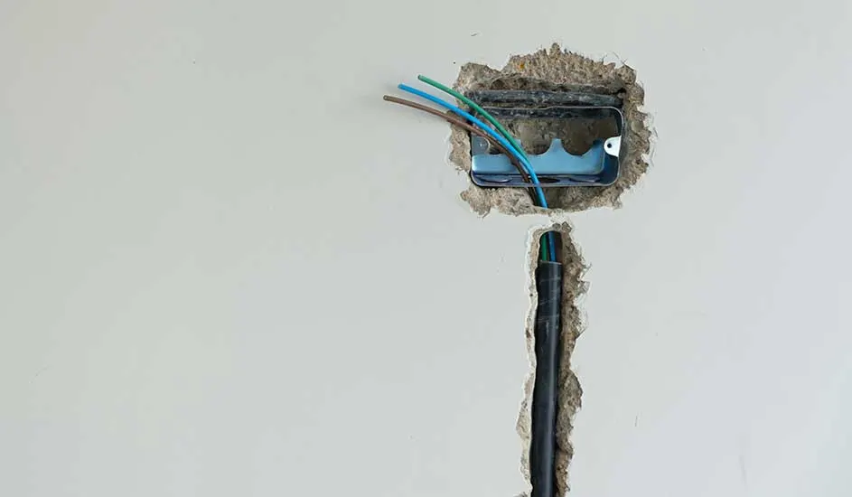 Een muur met een metalen elektriciteitsdoos en draden in blauw, bruin en groen. Een zwarte buis loopt verticaal onder de doos.