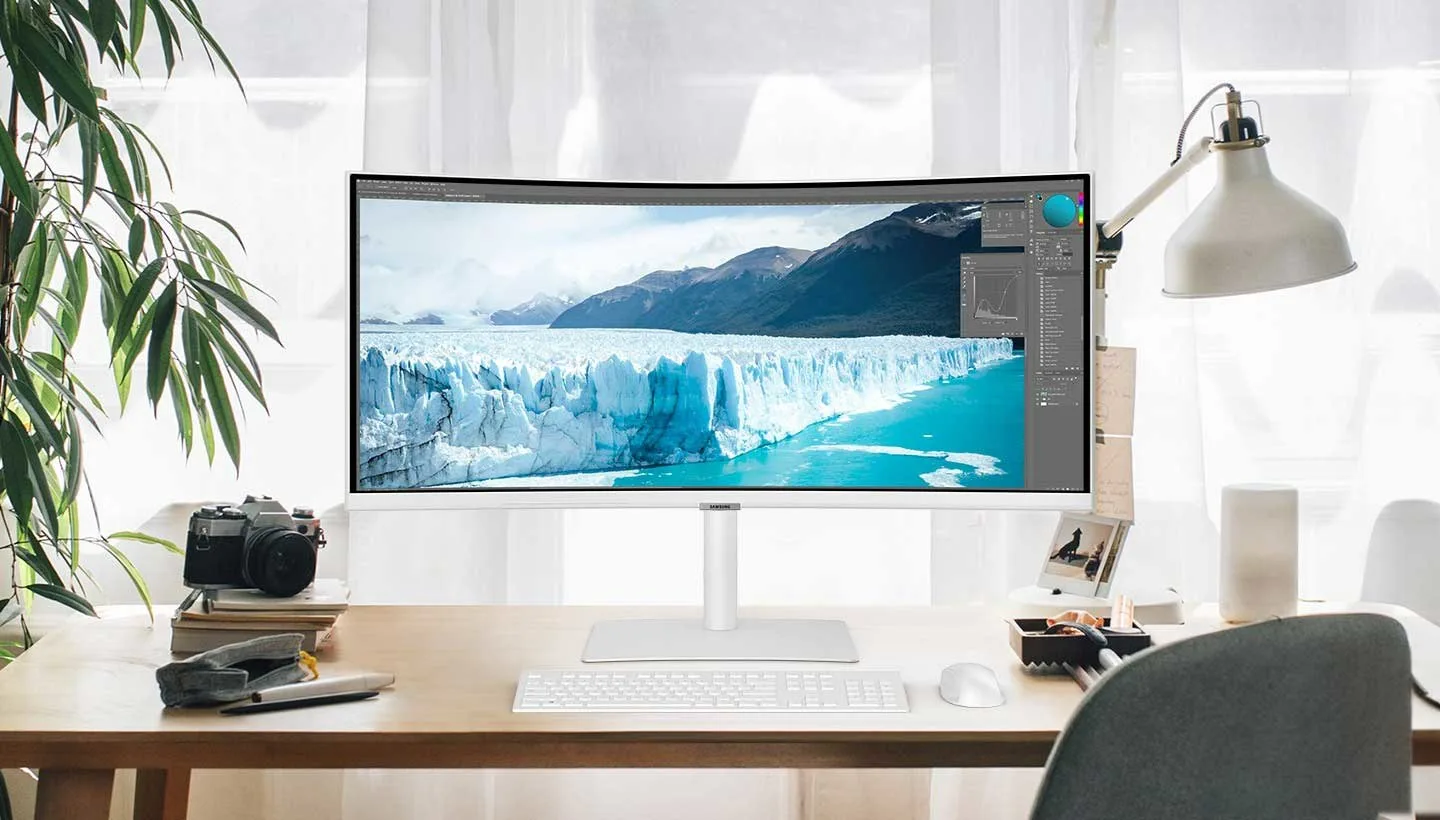 Een gebogen monitor toont een gletsjerlandschap op een houten bureau met een toetsenbord en een camera. Een plant en een lamp staan aan de zijkanten.