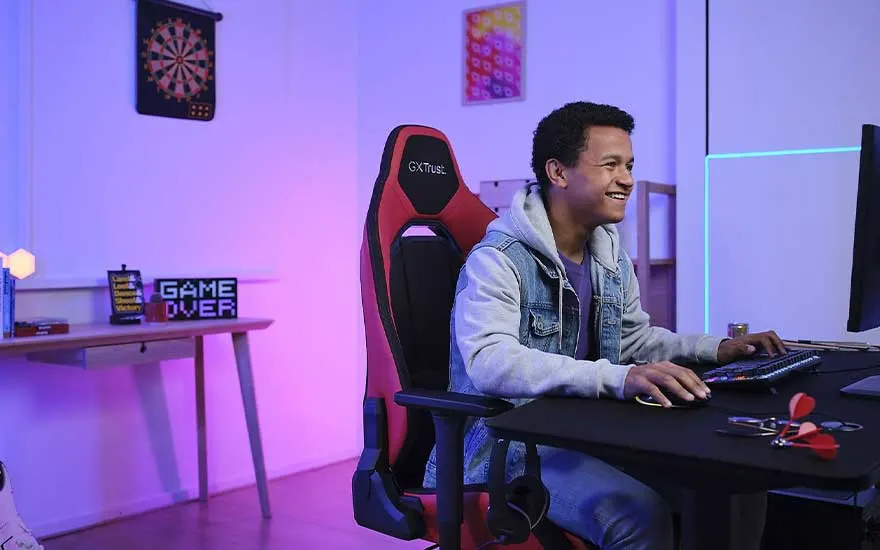 Een rode en zwarte gamingstoel staat voor een zwart bureau met een toetsenbord en muis. Op de achtergrond is een dartbord te zien.