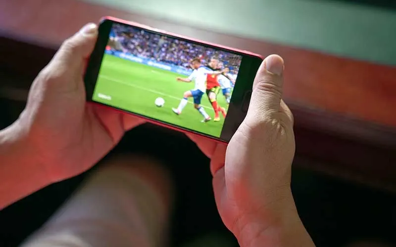 Iemand kijkt naar een voetbalwedstrijd op een rode smartphone. Het veld is groen en er is publiek op de achtergrond.