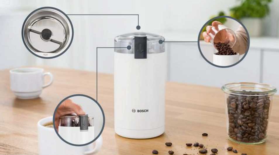 Een witte Bosch-koffiemolen staat op een houten tafel. Ernaast staan een kopje koffie en een pot met bonen; inzetbeelden tonen deksel en bediening.