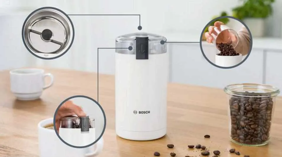 Een witte Bosch-koffiemolen staat op een houten tafel. Ernaast staan een kopje koffie en een pot met bonen; inzetbeelden tonen deksel en bediening.