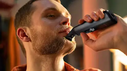 Un homme utilise une tondeuse à barbe noire. L'appareil affiche 100 % de batterie. Le fond est flou, avec des tons orange et blanc.