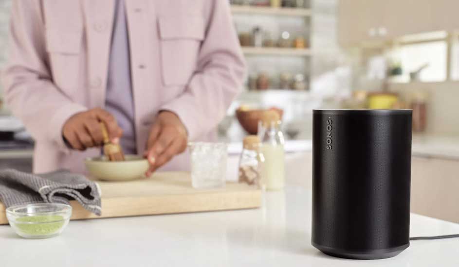 Sur le plan de travail se trouve une enceinte Sonos noire, tandis qu’une personne cuisine à l’arrière-plan dans une cuisine moderne.