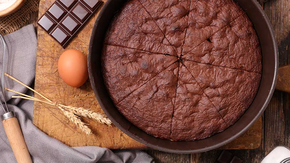 Un gâteau au chocolat coupé dans un moule rond, sur une planche avec un œuf, du chocolat, du blé et un fouet.