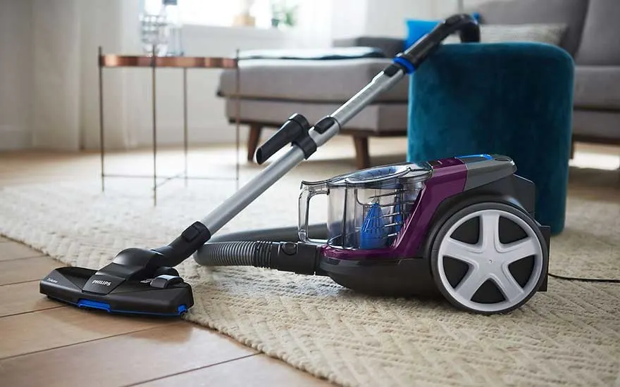 Een paarse Philips stofzuiger staat op een vloerkleed in een kamer met een blauwe poef en een tafel.