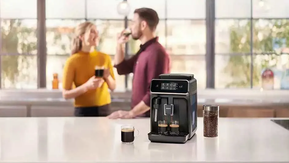 A black coffee machine sits on a white countertop, brewing into two small glasses. A glass jar of coffee beans sits nearby.