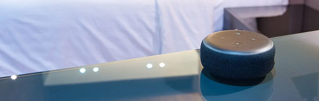 Een blauw-grijze slimme speaker staat op een glazen tafel, met een wit bed en nachtkastje vaag op de achtergrond.