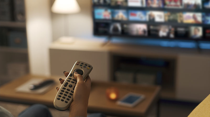 A hand holds a remote control in front of a TV displaying a menu of movies. A table and lamp are visible in the background.