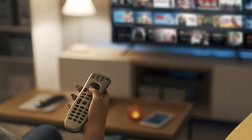 A hand holds a remote control in front of a TV displaying a menu of movies. A table and lamp are visible in the background.