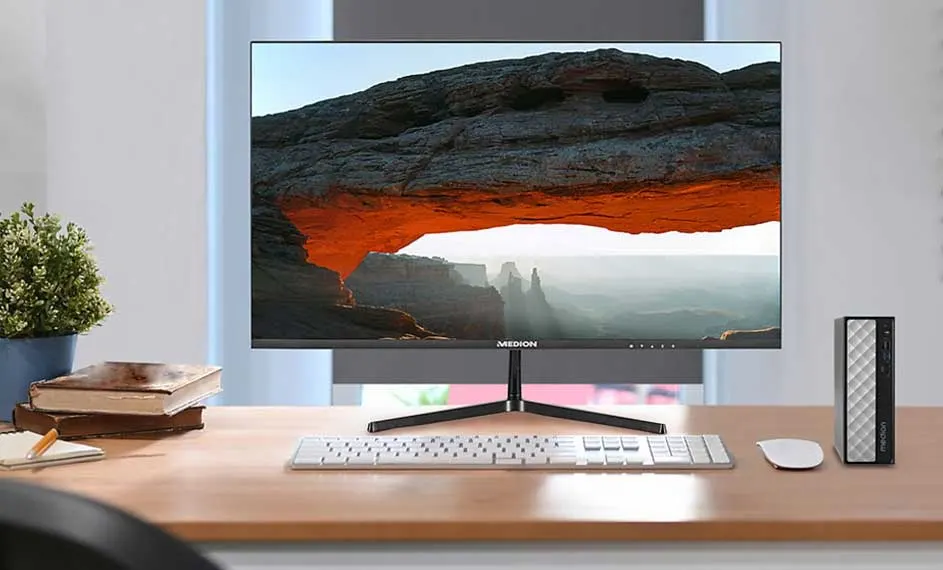 Bureau avec ordinateur Medion affichant un paysage rocheux, clavier blanc et mini-PC gris. Pot de plante et livres à gauche.