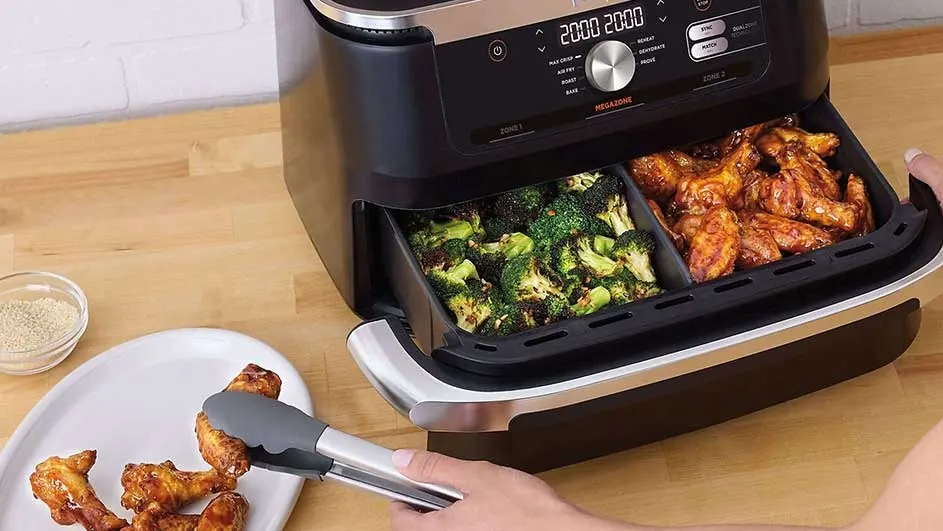 Air fryer with cooked broccoli and chicken wings in a divided drawer. Tongs grab wings from a plate.
