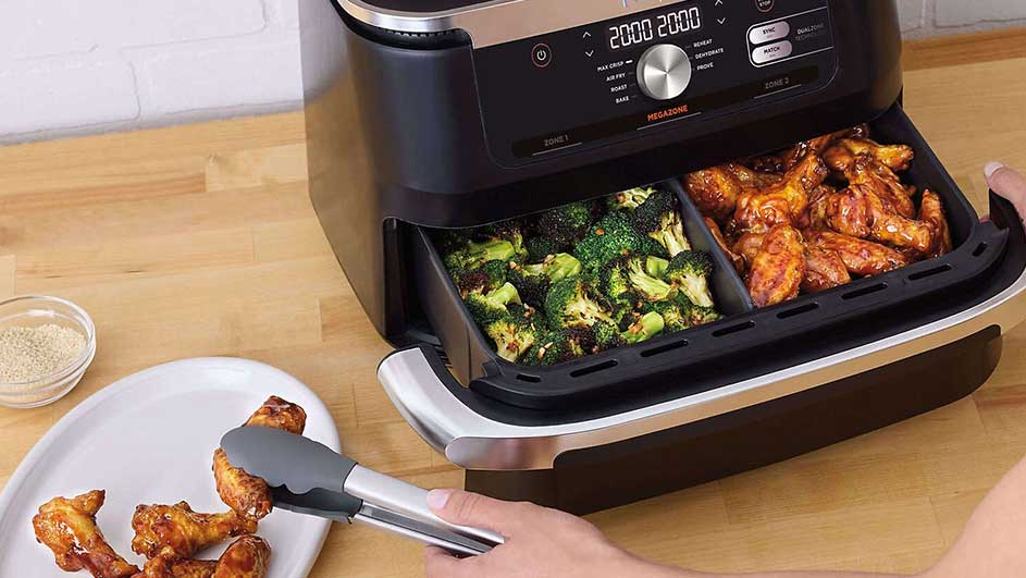 Een zwarte airfryer met twee lades gevuld met broccoli en kippenvleugels, op een houten tafel.