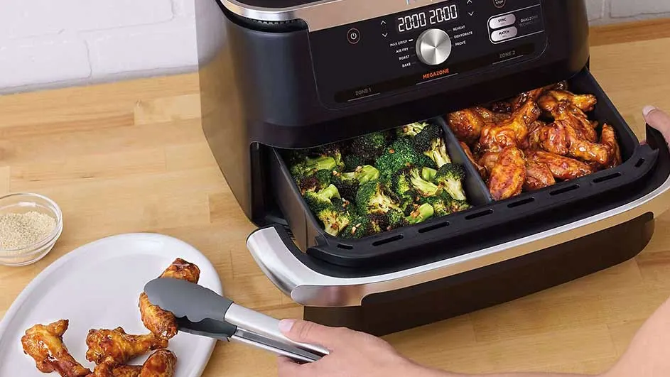 Een zwarte airfryer met twee lades gevuld met broccoli en kippenvleugels, op een houten tafel.