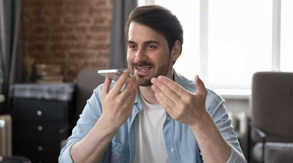 Un homme utilise un smartphone près de sa bouche, avec un mur de briques et une fenêtre en arrière-plan.