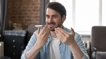 Un homme utilise un smartphone près de sa bouche, avec un mur de briques et une fenêtre en arrière-plan.