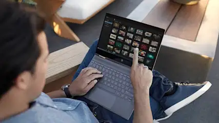 Een grijze laptop toont een scherm vol foto's op iemands schoot, met houten meubels op de achtergrond.