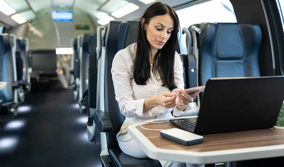 Femme voyage dans un train moderne, avec ordinateur portable et batterie externe sur la tablette, rechargeant son téléphone en lisant ses messages.