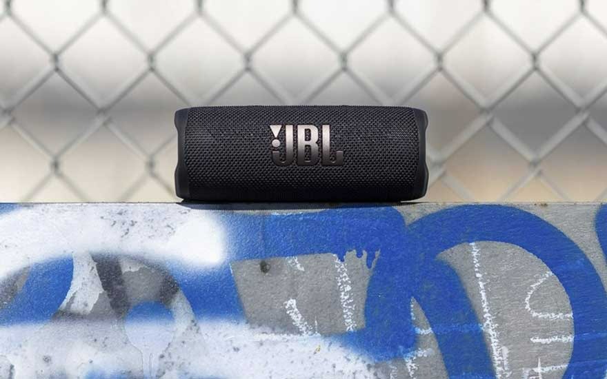 Haut-parleur JBL noir sur surface peinte en bleu et blanc, avec une clôture en métal en arrière-plan.