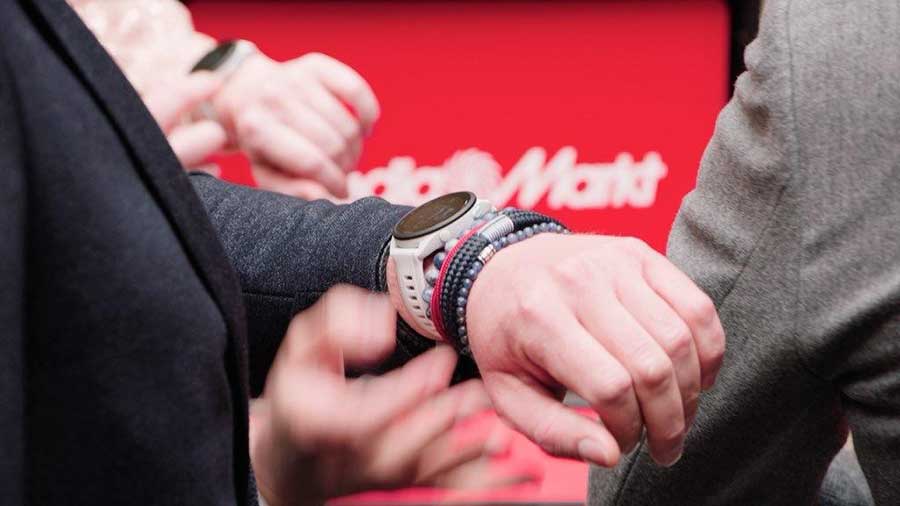 Een close-up van een pols met een witte smartwatch, kralenarmbanden en een rode achtergrond met een logo.