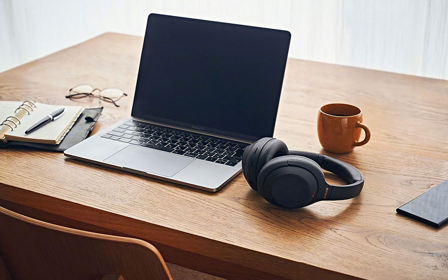 Een houten bureau met een laptop, zwarte hoofdtelefoon, notitieboek, bril, pen, bruine mok en smartphone.