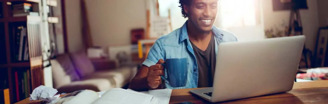 Een glimlachende man werkt thuis aan een laptop, houdt een blauwe mok vast en heeft papieren en boeken op de houten tafel liggen.